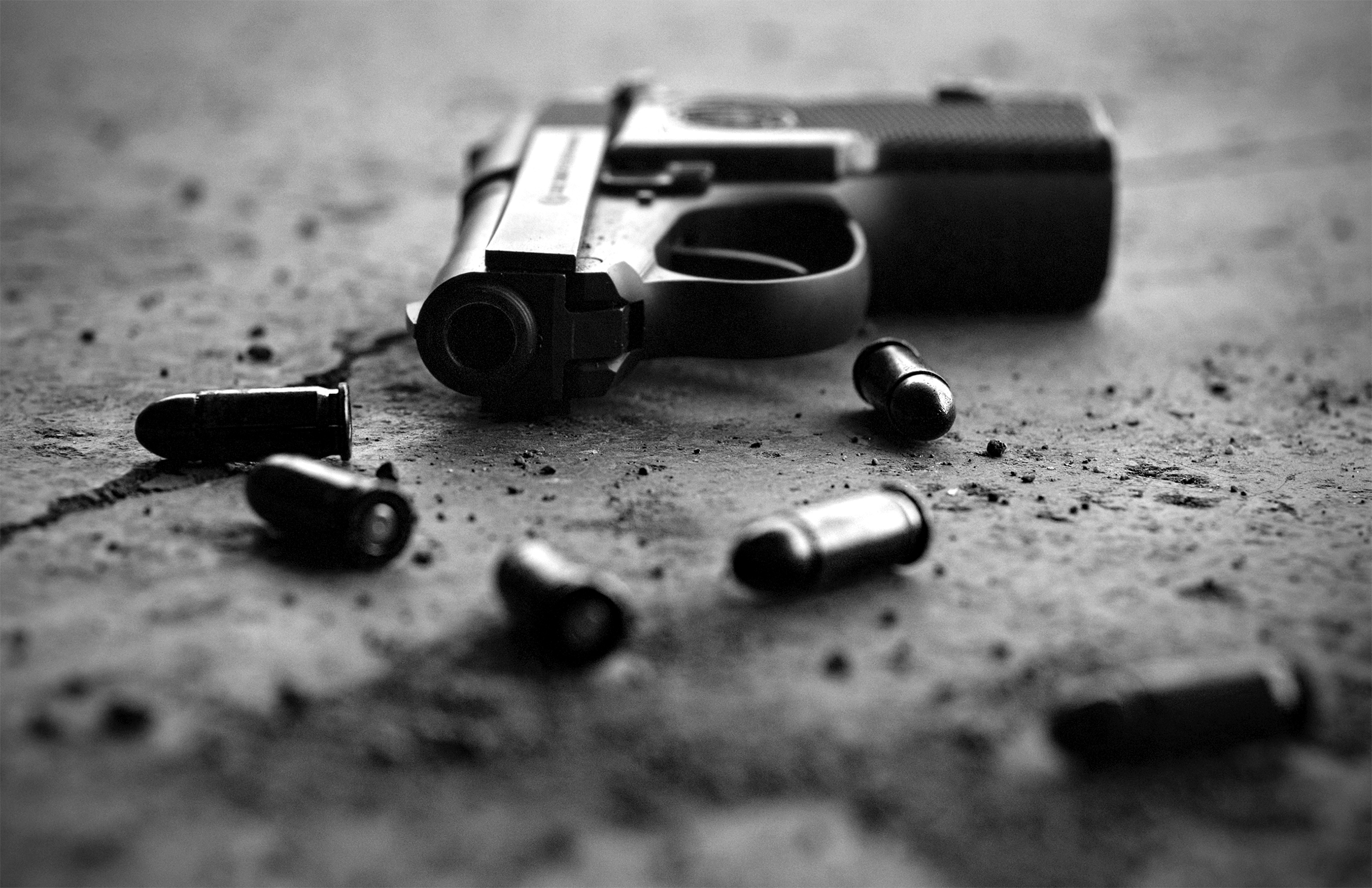 A close-up of a pistol and six bullets lying on the ground. The barrel of the pistol is pointed toward the camera.