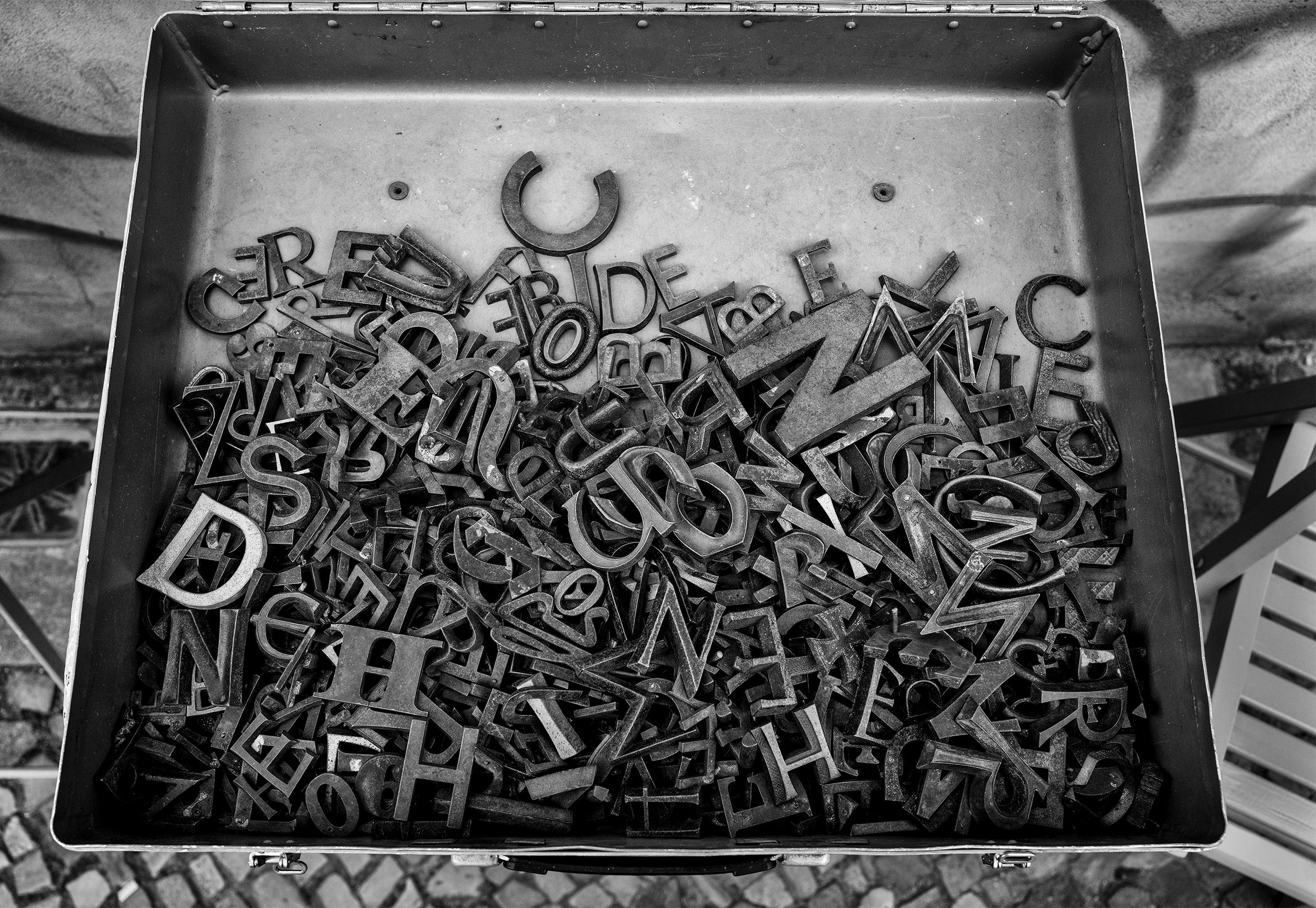 The bottom part of a hinged metal box half full of metal letters of various sizes.