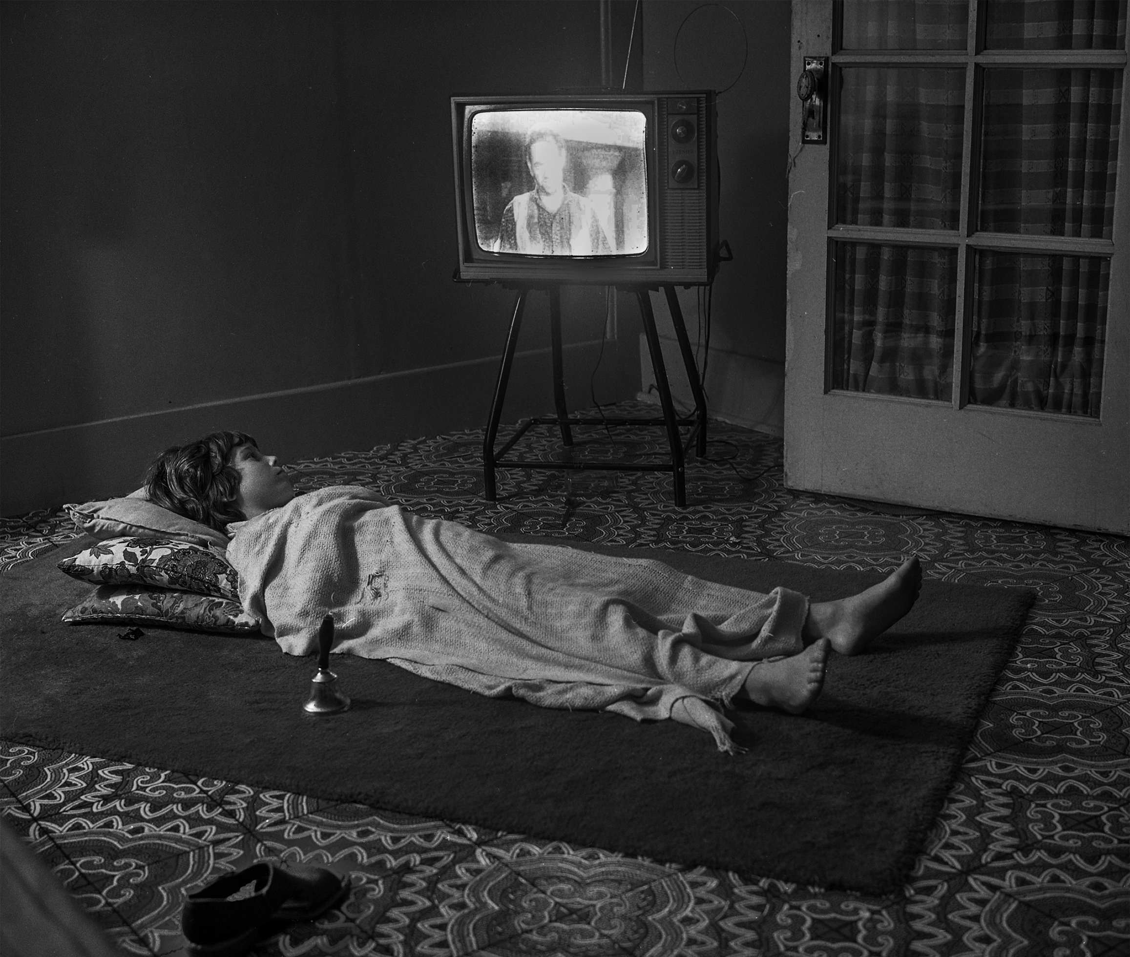 A young boy in the 1970s is flat on his back on the floor atop an area rug on top of carpeting with three pillows under his head and covered with a blanket in a room with an old-fashioned television. There is a long-handled bell near him on the rug.