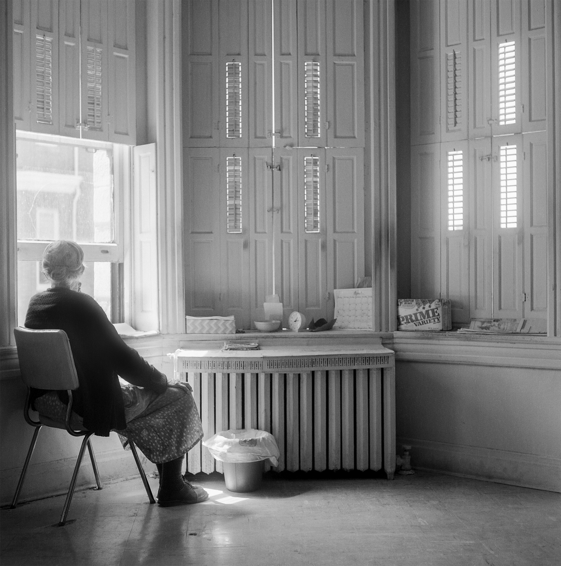 An old woman, with her hair in a bun and wearing a dress and sweater, sits in a chair and looks out of the window. The room has a steam radiator air vent, tall windows with interior shutters, a small wastebasket, and personal items on the windowsill.