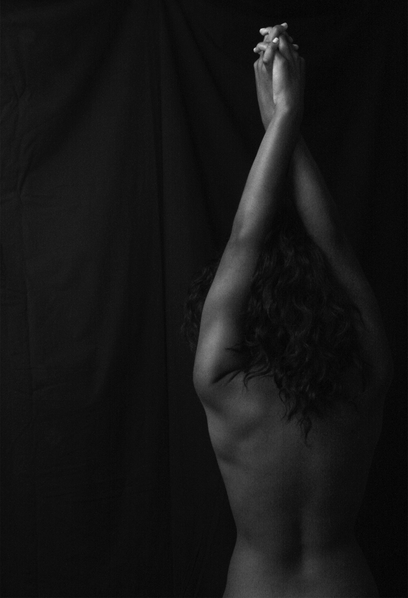 A nude woman with long, curly hair is seen from the back and above the waist standing with her arms fully extended above her head with her hands crossed and fingers intertwined.