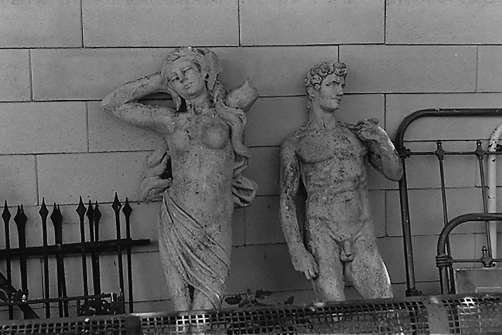 Two statues propped against a wall in a storage area amid decorative metal fencing. A statue of a mostly nude female on the left and a replica of Michelangelo’s David on the right.