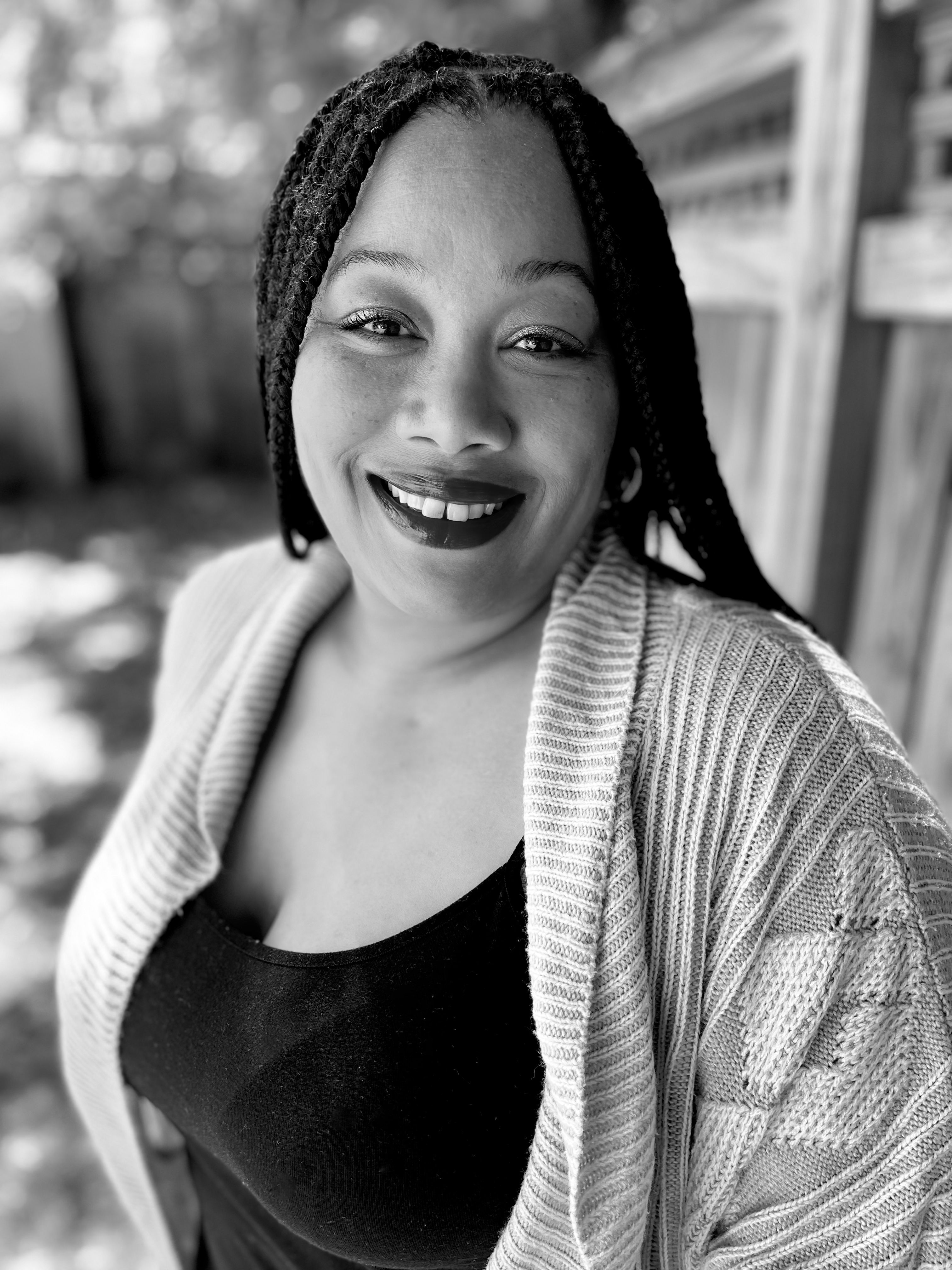 A Black woman with dark braided hair stands outside the Sun's office in front of a wooden fence. She smiles at the camera and wears a dark top under a light-colored sweater.